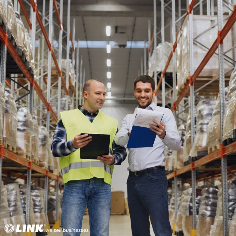 Two men in a warehouse reviewing paperwork on clipboards, amid tall racks of pallet-stacked inventory.
