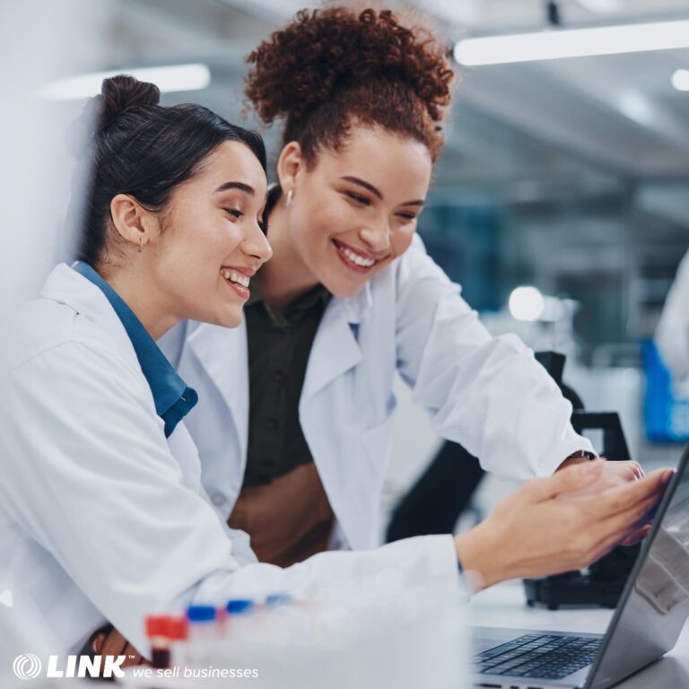 Two lab-coated individuals smiling and reviewing a laptop together, with vials in the foreground.